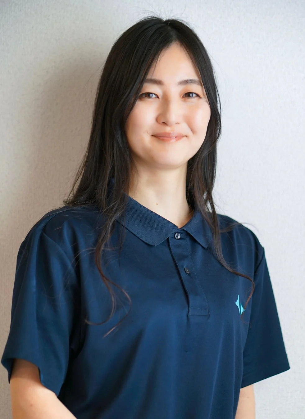 紺色のポロシャツを着た若いアジア人女性が、白い壁を背景に笑顔で写っています。
