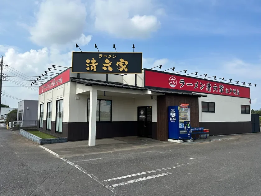 青空の下、ラーメン店「清六家 江戸崎店」が建つ風景です。店舗の看板には店名が大きく表示され、入口付近にはBOSS COFFEEの自動販売機が設置されています。周辺には広々とした駐車場があり、郊外の店舗であることが伺えます。