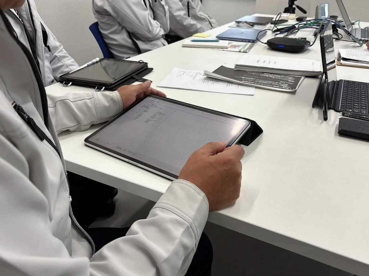 作業着を着た人物が会議中にタブレットを操作し、画面上のデータを確認している様子。会議室のテーブルには複数のデバイスや資料が置かれ、ビジネスシーンでの情報共有や打ち合わせの風景を捉えている。