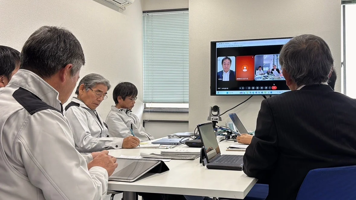 会議室で複数人がハイブリッド会議に参加している様子です。一部の参加者は対面で、別の参加者は大型スクリーンに映し出されたビデオ通話を通じてリモートで参加しています。