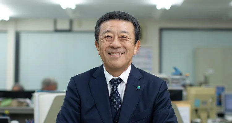 オフィスでネイビーのスーツを着用した日本人の中年男性が、カメラに向かって明るく笑顔を見せているポートレート写真です。親しみやすく、プロフェッショナルな印象を与えます。