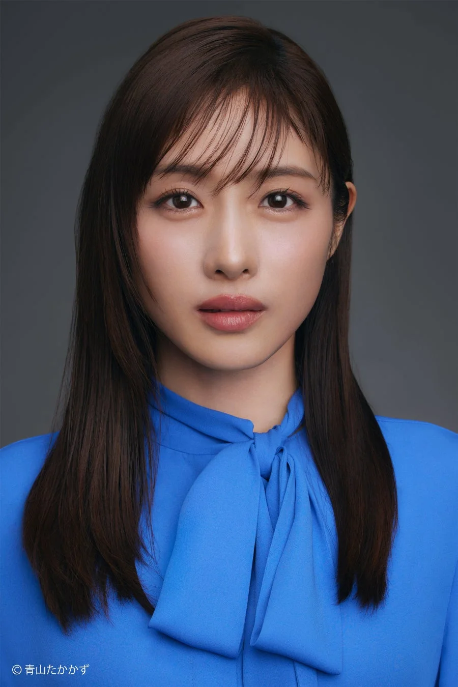 青いブラウスを着用したロングヘアの日本人女性のポートレート。カメラをまっすぐ見つめており、落ち着いた表情が印象的です。背景はシンプルなグレーです。