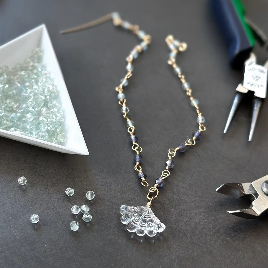 扇形のペンダントトップが特徴的なハンドメイドネックレスの制作風景です。青や紫系のビーズが連なるネックレスの隣には、制作途中のビーズや工具が置かれ、手作りの温かみを感じさせます。