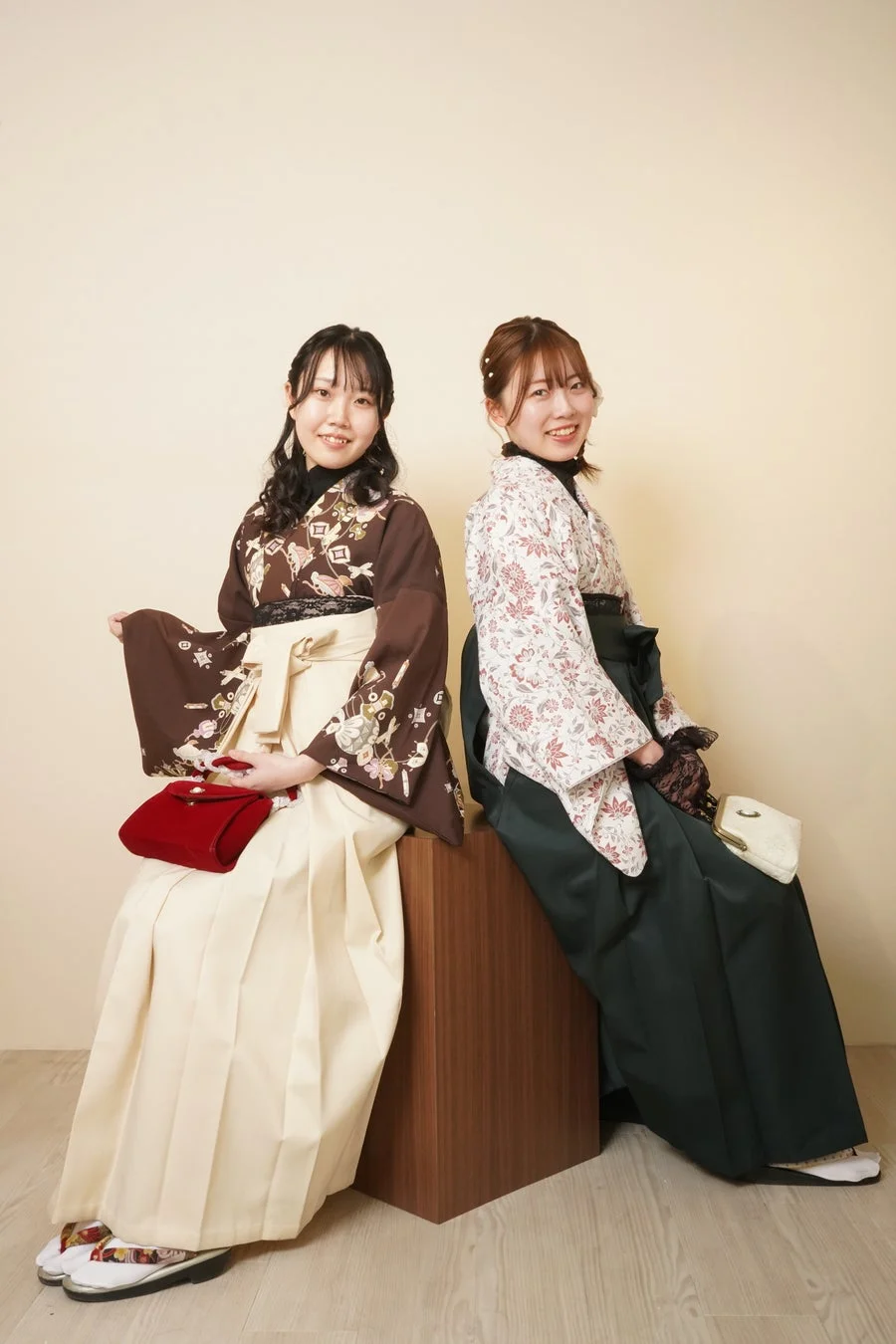 袴姿の若い女性二人が笑顔で並んで座っているポートレートです。茶色の着物にベージュの袴、白地の花柄着物に濃緑の袴をそれぞれ着用し、卒業式のような華やかな雰囲気が伝わります。