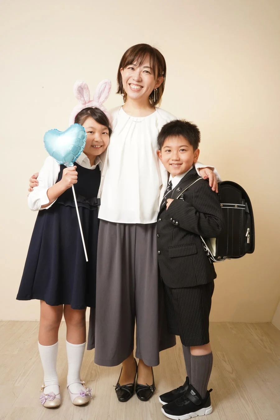 笑顔の母親と、スーツ姿でランドセルを背負った男の子、ドレスとうさ耳、ハートの風船を持った女の子が並んで写っている記念写真。入学式などのお祝いの場面を思わせる温かい雰囲気の家族写真です。