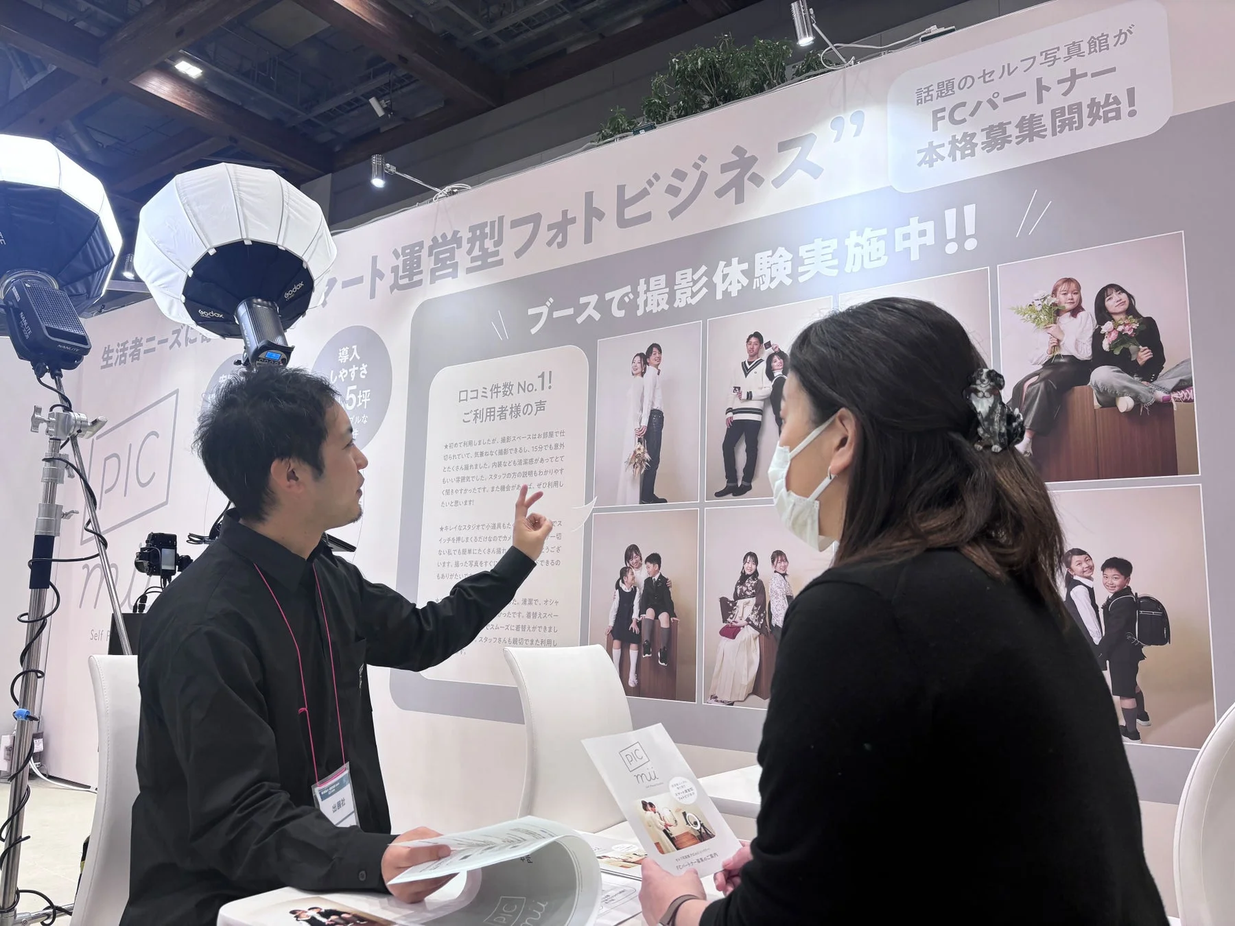 展示会ブースで、セルフ写真館「PIC miu」のFCパートナー募集が行われている様子。男性スタッフが女性来場者にサービス内容を説明しており、導入のしやすさや顧客の口コミ、撮影体験が紹介されている。