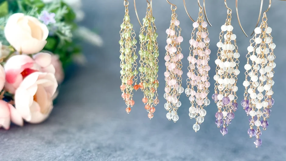 様々な色の天然石ビーズが連なった、繊細なロングピアスが複数並べられています。背景にはぼかされた花が写っており、上品で可愛らしい雰囲気のアクセサリーコレクションです。