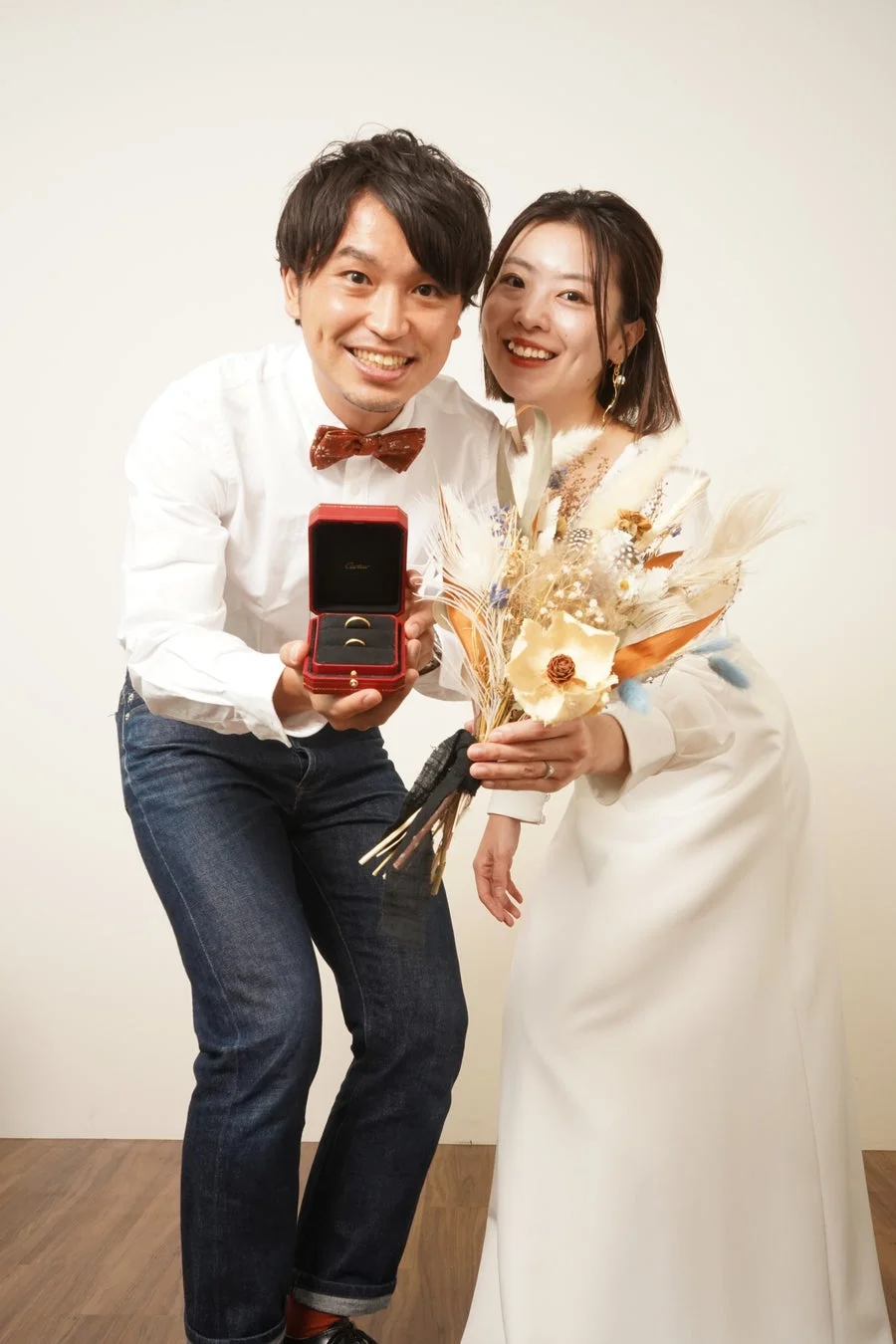 幸せそうなカップルがカメラに向かって笑顔を見せています。男性は赤いCartierのリングボックスを開けており、中には結婚指輪らしき2つの指輪が見えます。女性は白いドレスを着て、ドライフラワーのブーケを持っています。