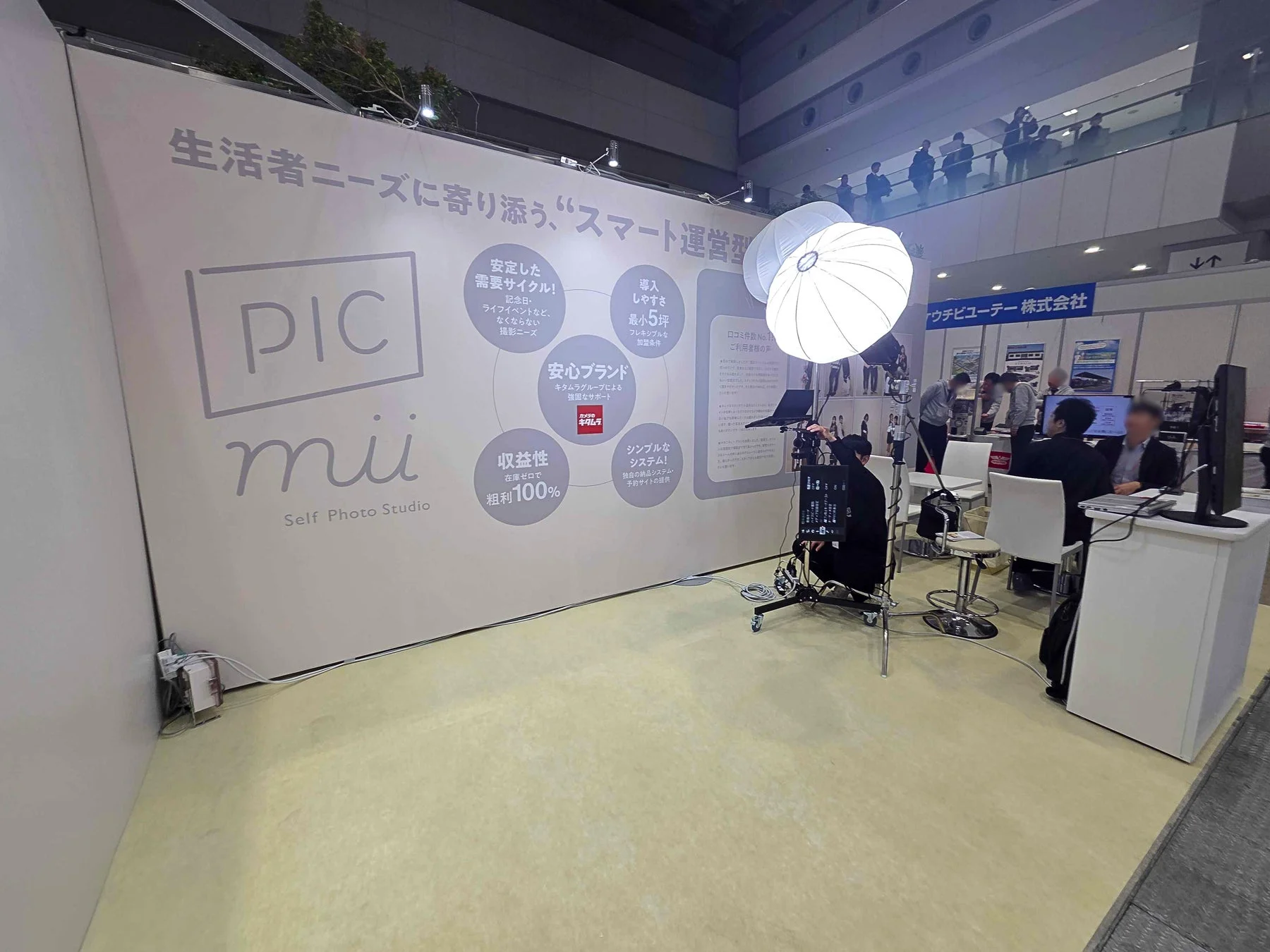 展示会ブースで「PIC mii Self Photo Studio」のビジネスソリューションが紹介されています。壁面には、消費者ニーズに寄り添うスマート運営、安定した需要、導入のしやすさ、安心ブランド（キタムラグループ）、高収益性、シンプルなシステムといった特徴が掲示されており、来場者とスタッフが商談している様子が伺えます。