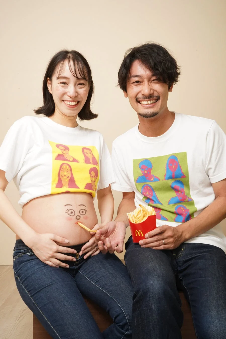 笑顔の夫婦がマタニティフォトを撮影しています。女性のお腹には顔が描かれており、男性がマクドナルドのフライドポテトをその顔に食べさせているユニークで楽しい瞬間です。