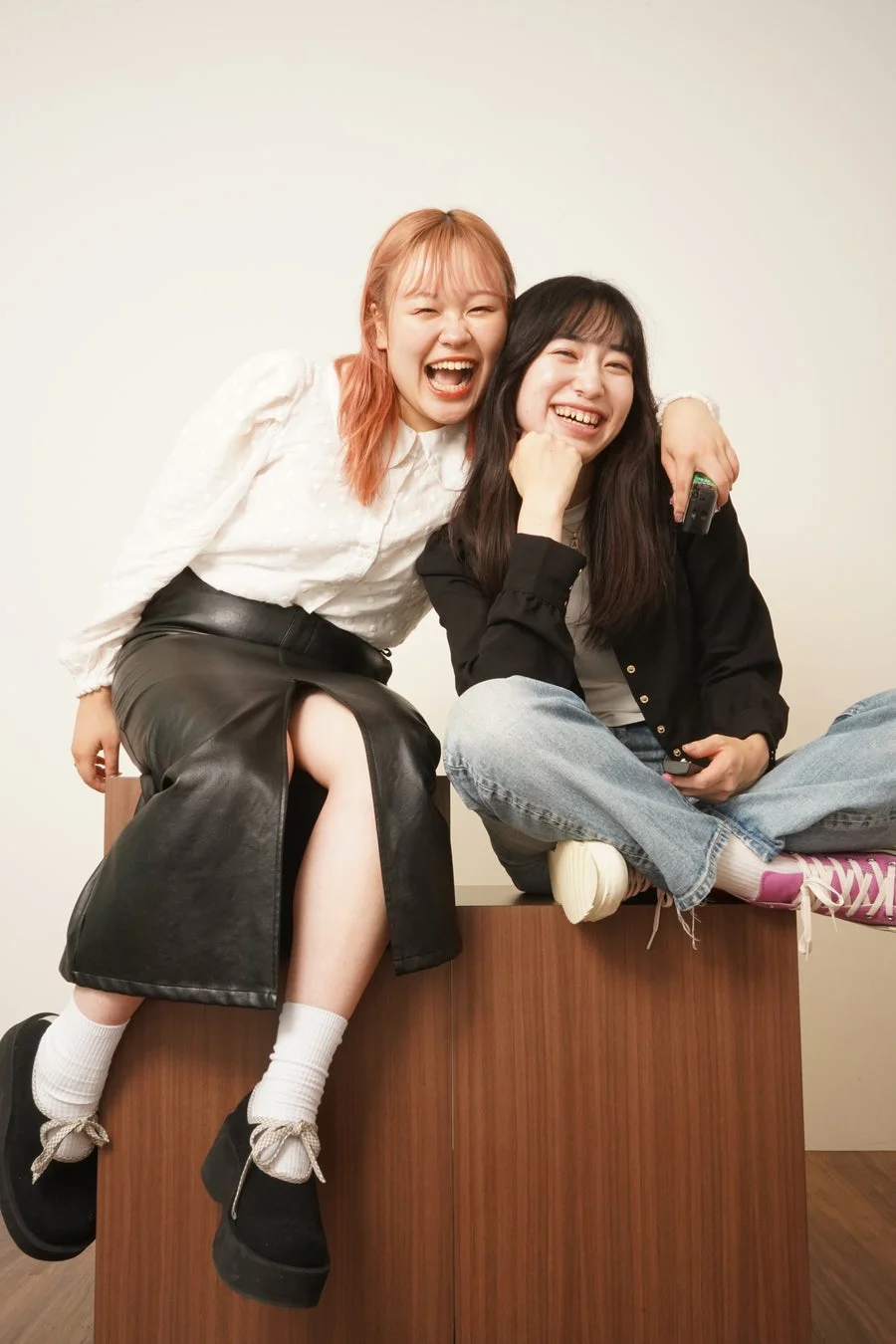 白い背景の前で、二人の若い女性が笑顔で楽しそうに座っています。一人はピンク色の髪で白いブラウスと黒いスカート、もう一人は黒髪で黒いジャケットとジーンズを着用しており、親密な友情を感じさせる一枚です。