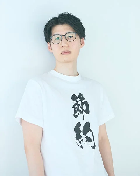 白いTシャツと眼鏡をかけた若い男性が、白い背景の前で正面を向いています。Tシャツには「節約」という文字が書かれています。