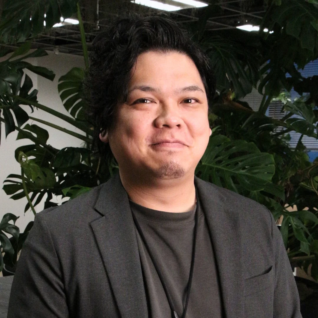 緑豊かな植物を背景に、ダークグレーのジャケットを着たアジア系の男性