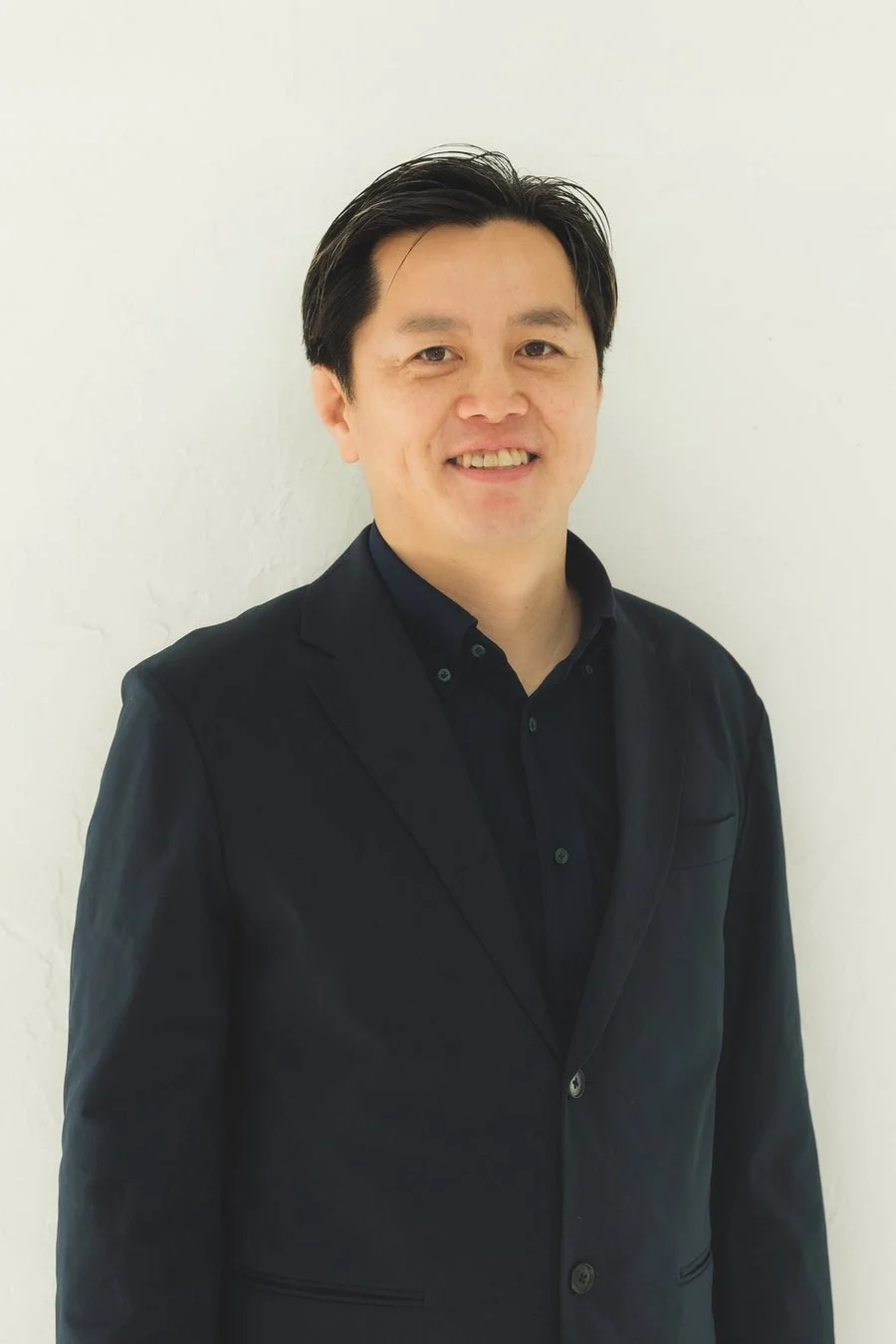 白い背景の前で、ダークスーツとシャツを着用し、笑顔でカメラを見つめるアジア人男性のポートレート