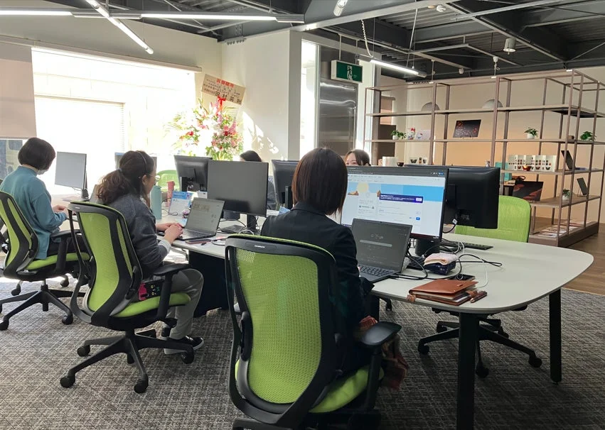 明るく開放的なオフィスで、数人のスタッフが各自のデスクでパソコンに向かい作業している様子です。奥には「祝御就任」と書かれたお祝いの札が見えます。緑色の椅子が印象的な、モダンな職場環境です。