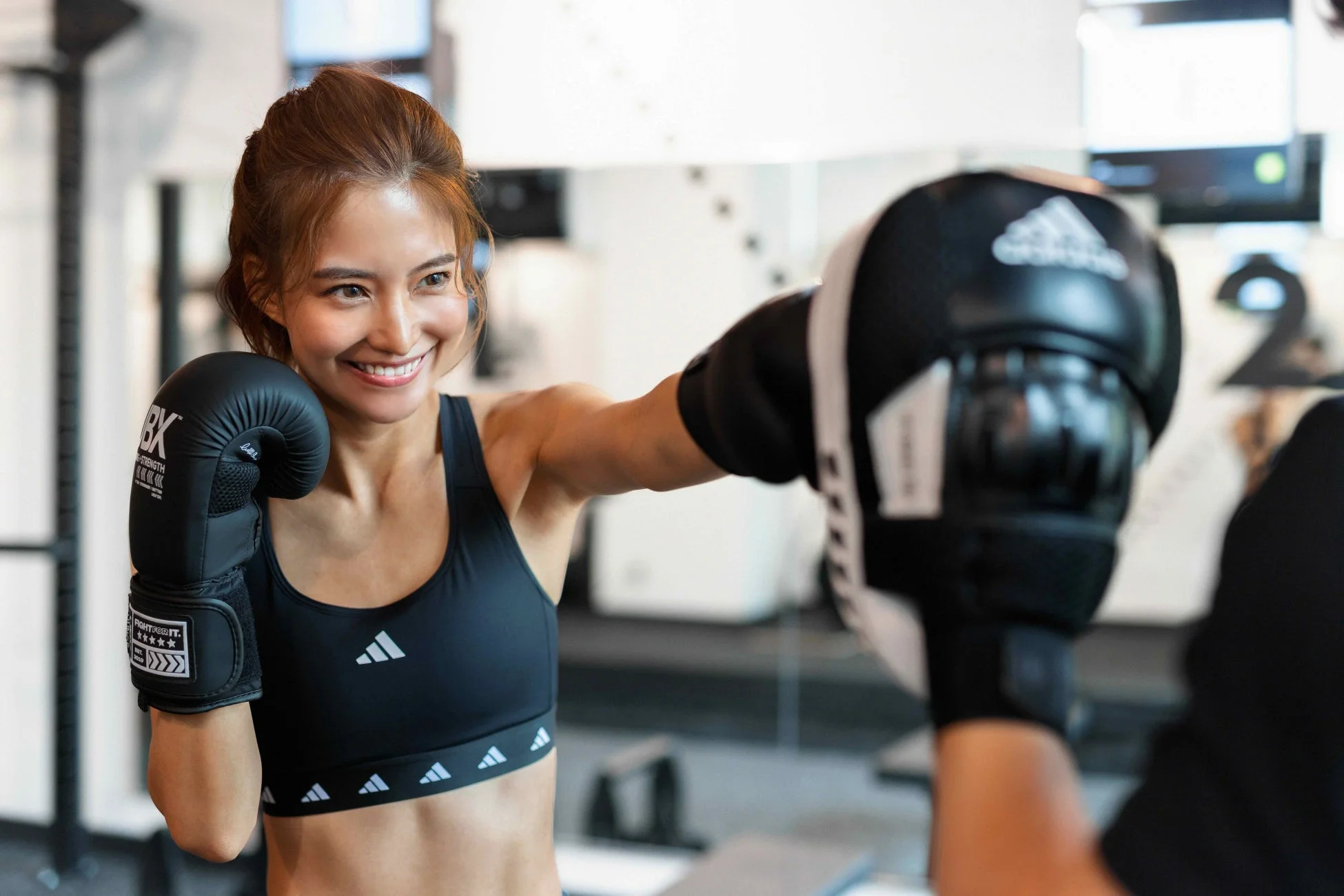 ジムで女性がボクシンググローブとスポーツブラを着用し、笑顔でミット打ちをしている瞬間