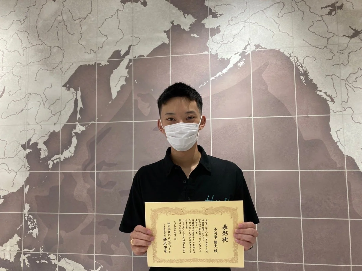 世界地図を背景に、マスクを着用した男性が表彰状を手にしている
