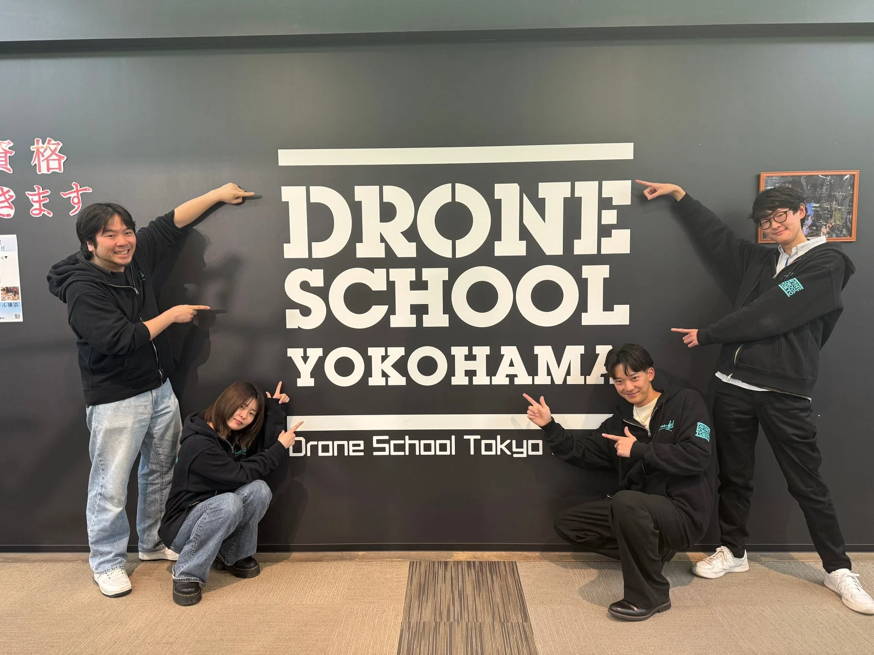 黒い壁の「DRONE SCHOOL YOKOHAMA」ロゴの前で、4人の男女が笑顔でポーズをとっている集合写真
