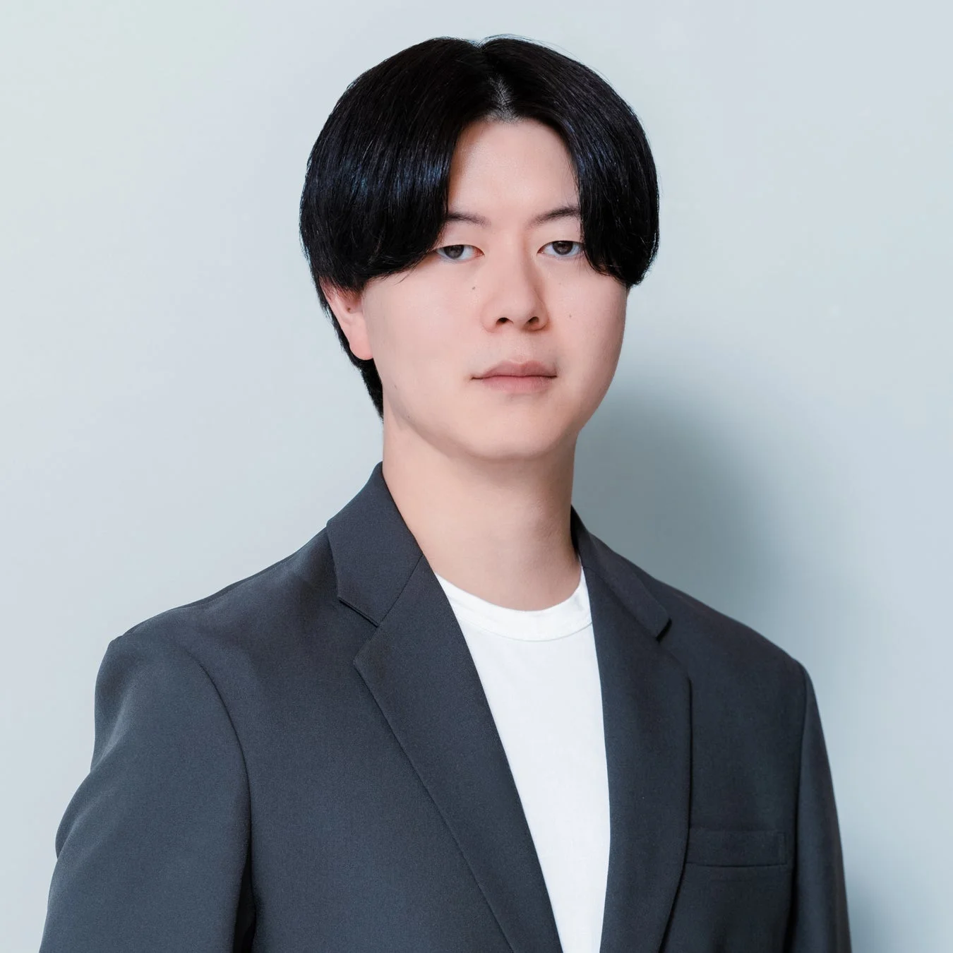 明るい背景の前で、ダークグレーのジャケットと白いTシャツを着用した黒髪の若いアジア人男性が、カメラをまっすぐ見つめている上半身のポートレートです。