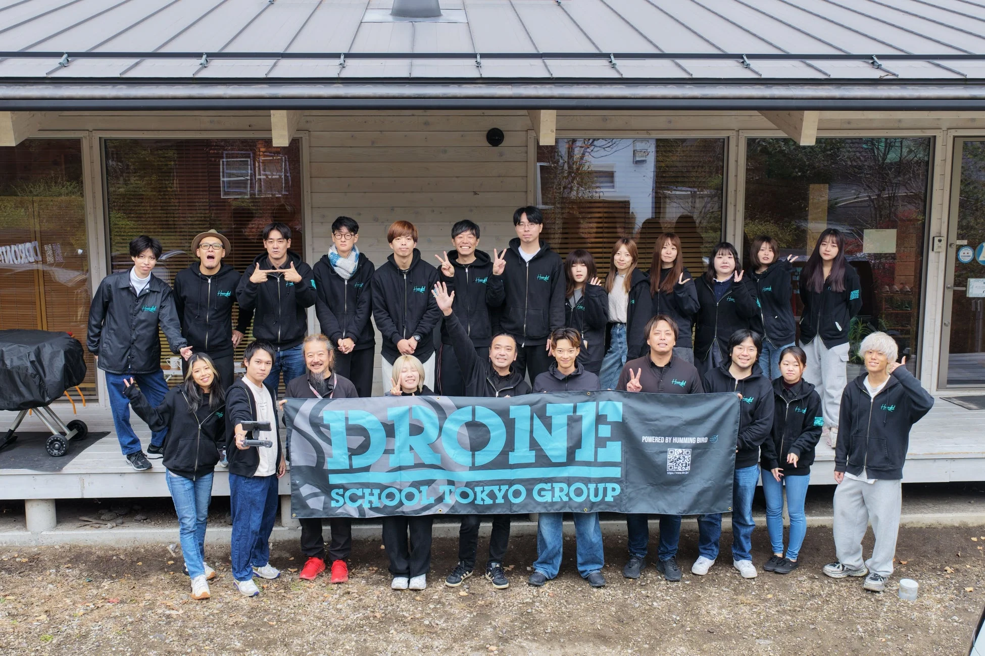 「DRONE SCHOOL TOKYO GROUP」と書かれたバナーを囲んで、多くの男女が建物の前で笑顔でポーズをとっている集合写真