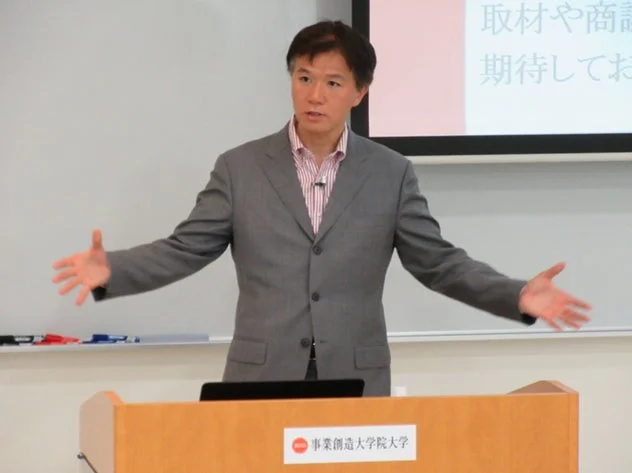 スーツを着た男性が壇上でプレゼンテーションを行っています。背景にはプロジェクターで映し出された資料が見え、演台には「事業創造大学院大学」のロゴがあります。