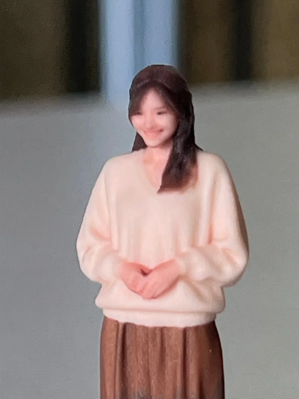 ピンク色のVネックトップスと茶色のプリーツスカートを着用し、笑顔で両手を前で組んでいる若い女性の3Dフィギュア