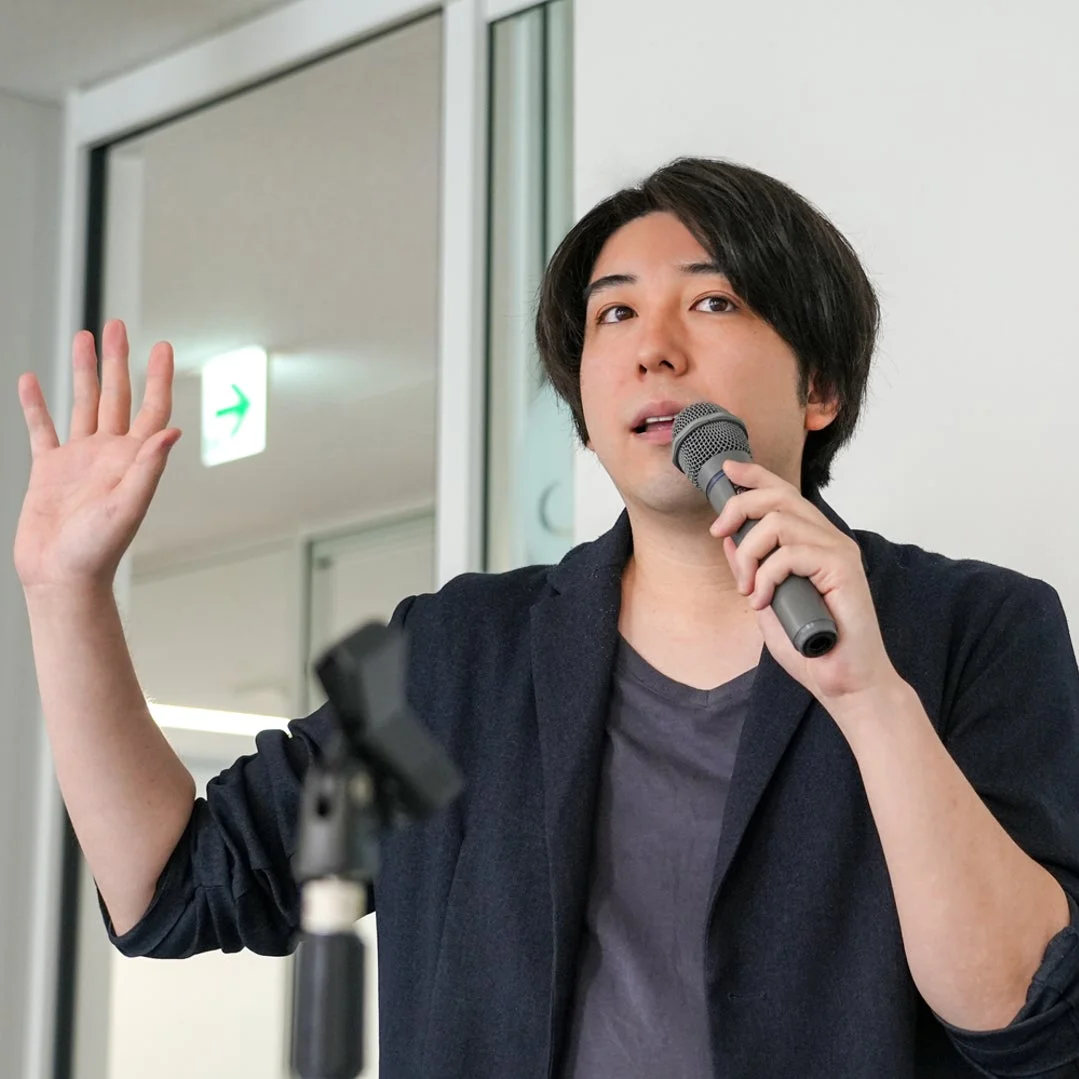 マイクを持って講演中の男性の姿。片手を上げてジェスチャーを交えながら話しており、プレゼンテーションか会議の場面と推測される。背景には右向きの緑色の矢印サインが見える。