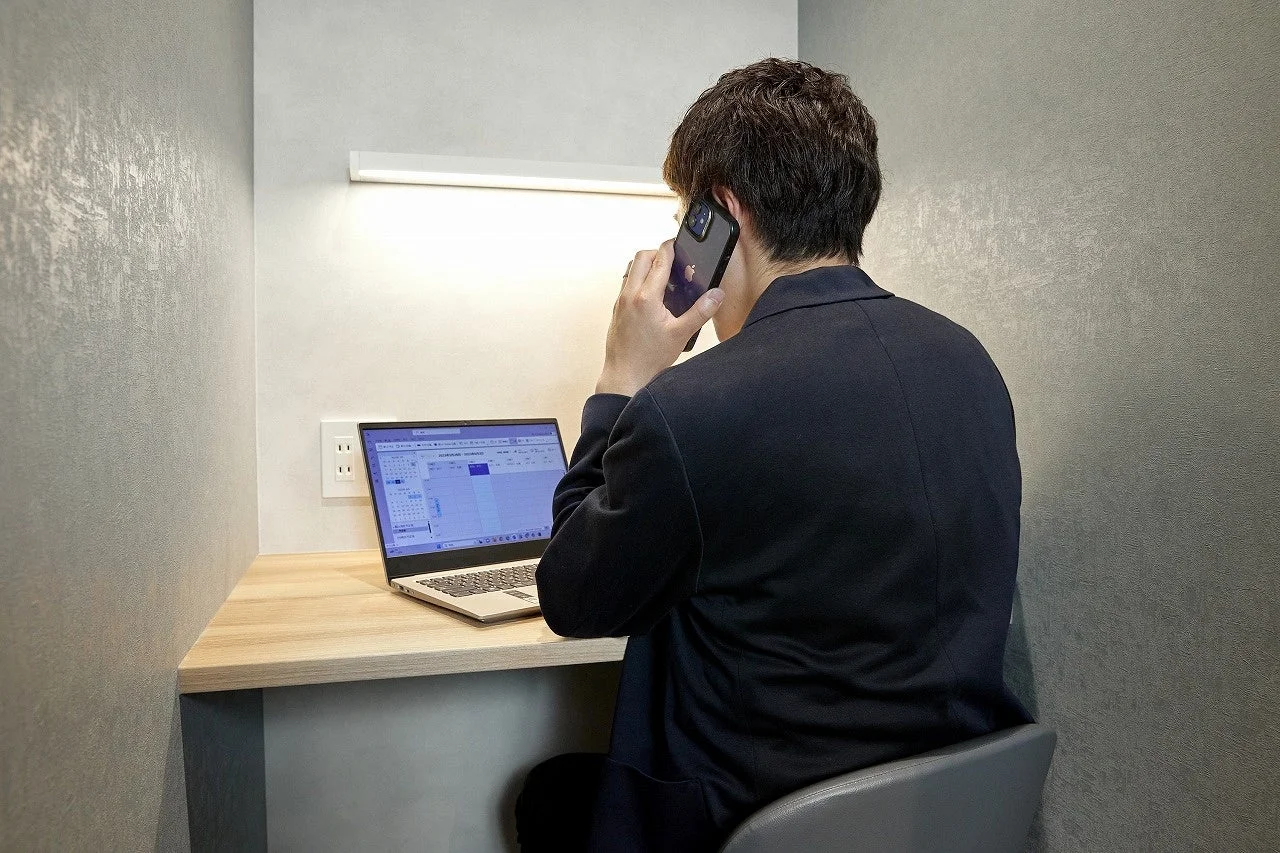 個室ブースで仕事をする男性の後ろ姿を捉えた画像です。男性はスマートフォンで通話しながら、ノートパソコンの画面を見ています。集中して仕事に取り組む様子が伺えます。