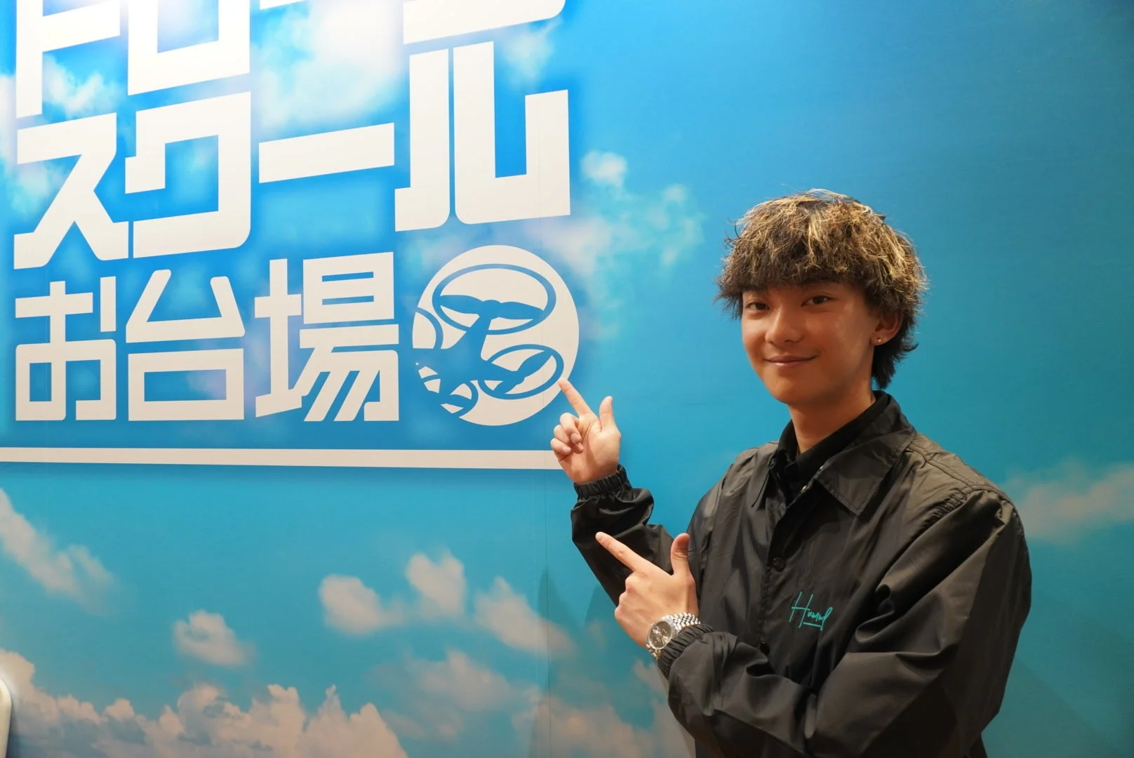 男性がドローンスクールお台場の看板とドローンを指さして笑顔を見せている