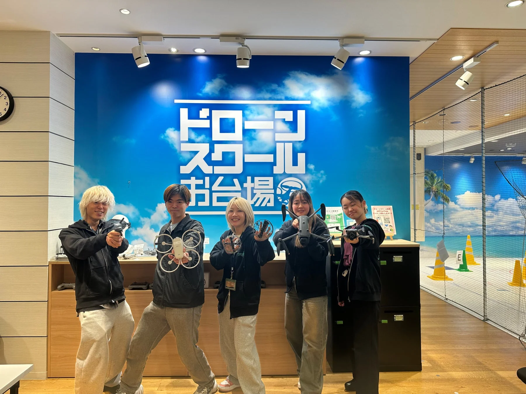 「ドローンスクールお台場」の屋内施設で、5人の若者がドローンを持って楽しそうにポーズをとっている