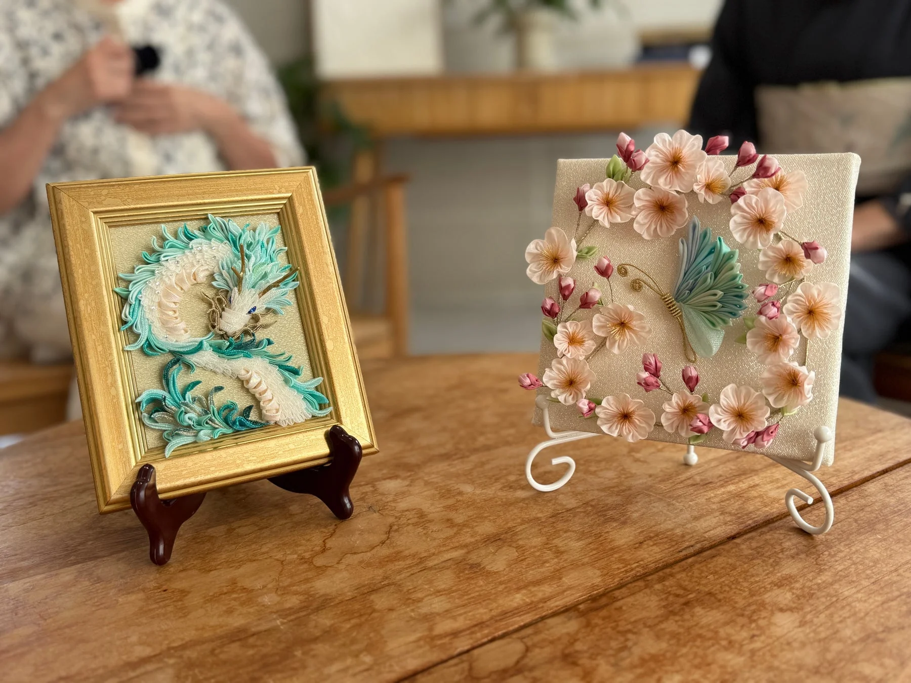 木製のテーブルに置かれた2つの美しい手芸品。左は金色の額に入った青と白の龍のアート、右はピンクの花と蝶が飾られた作品です。