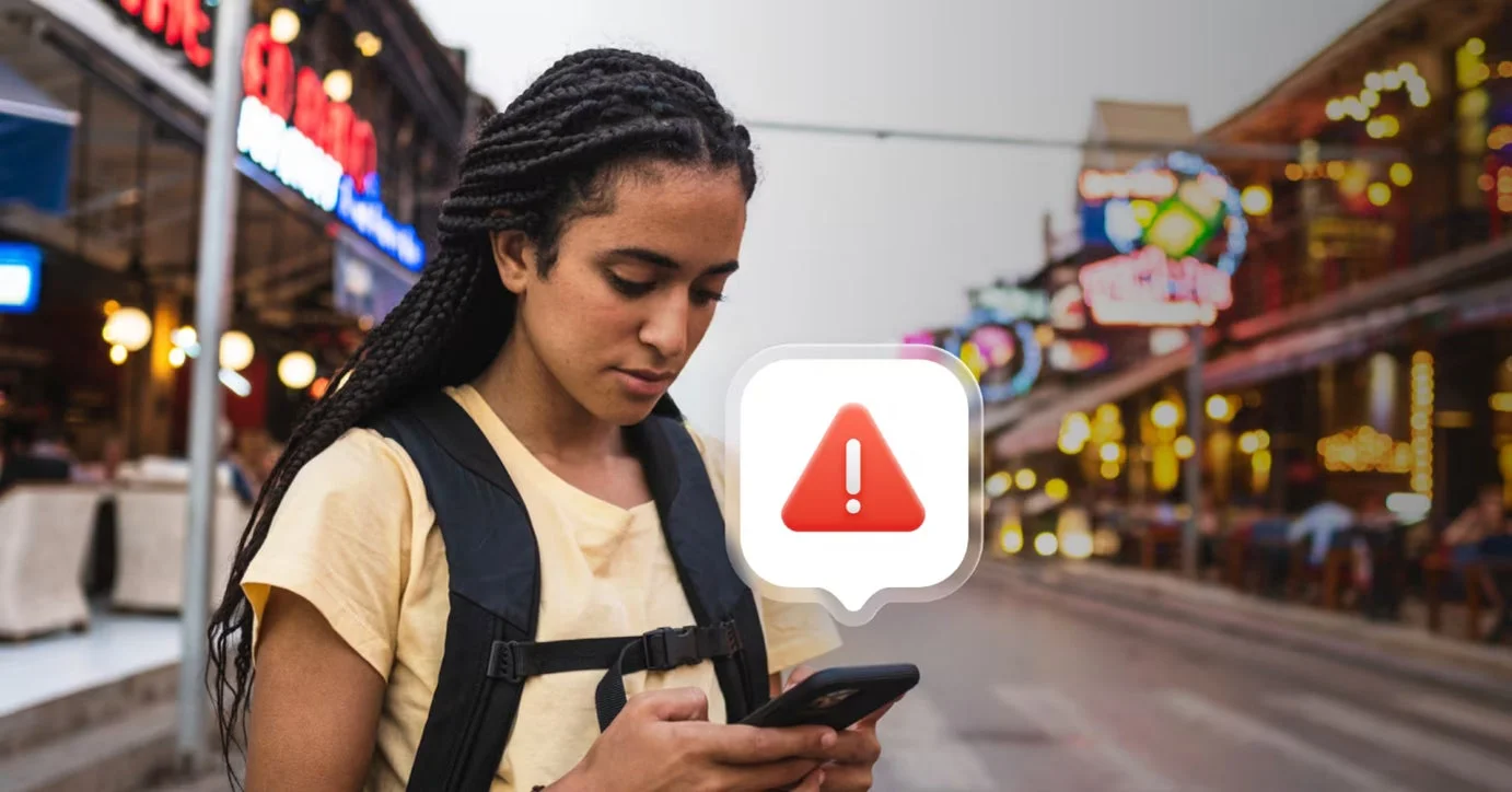 夜の賑やかなストリートで、一人の女性がスマートフォンを操作しています。画面の上には警告を示す赤い感嘆符のアイコンが表示されており、何らかの問題が発生していることを示唆しています。