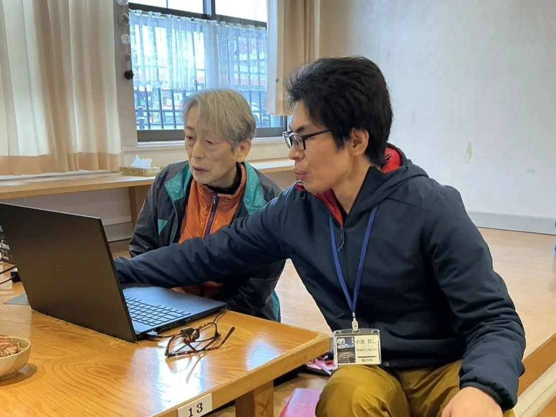 一人の高齢者と若い男性がノートパソコンを見ながら話している場面です。若い男性が高齢者にパソコンの使い方を教えているようで、デジタル機器の学習会やサポートの様子を捉えた写真です。