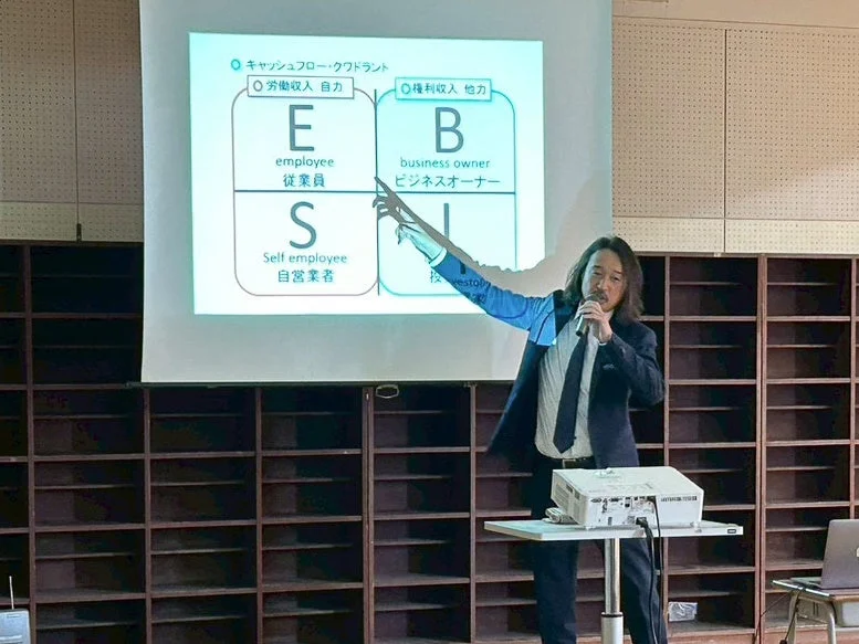 スーツを着た男性が、キャッシュフロー・クワドラントの図を映したスクリーンを指しながら、聴衆に向けて講演している様子