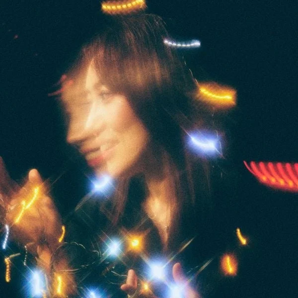 暗い背景にぼかされた女性の顔が写っており、その周りには多数の光の筋やフレアが輝いています