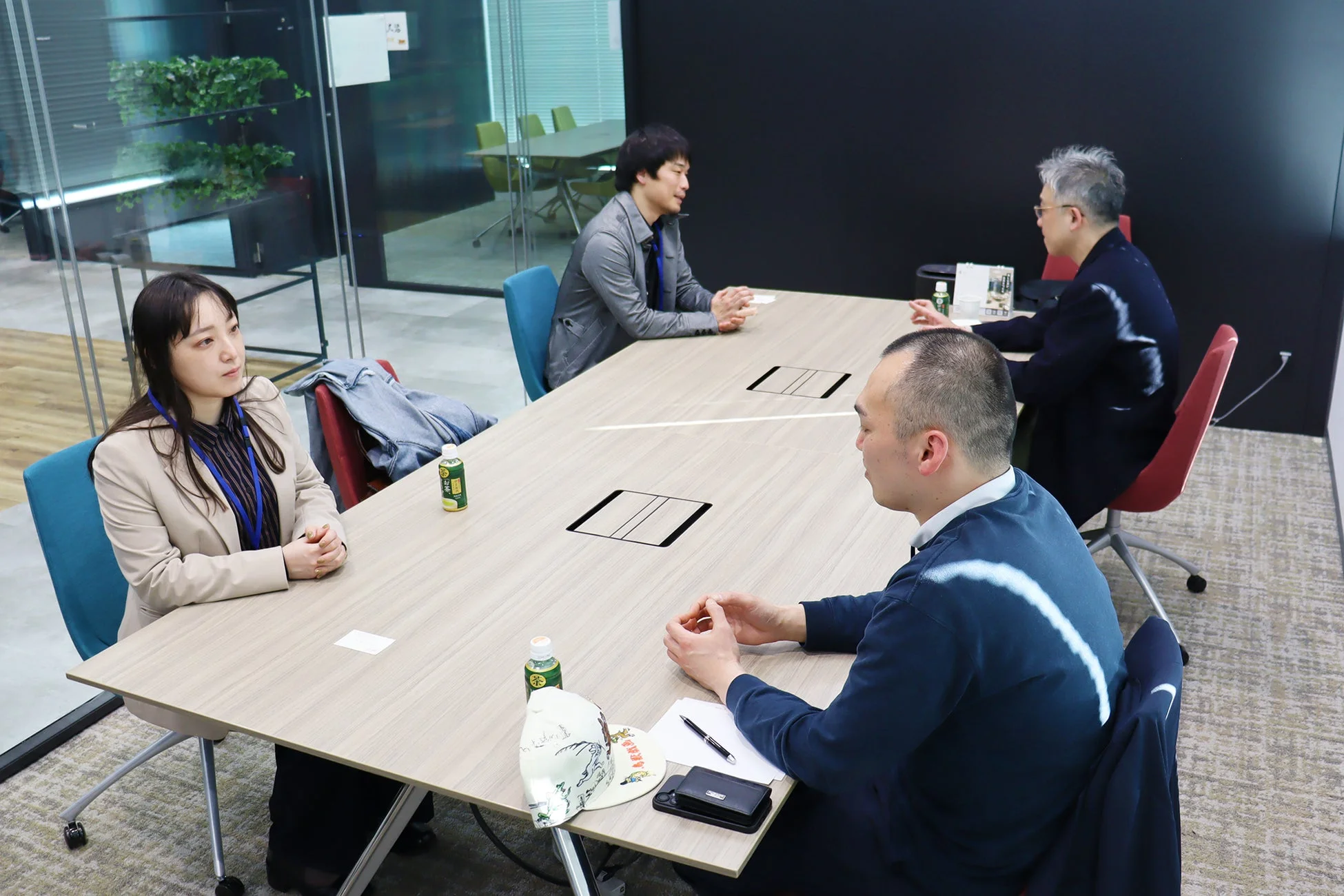 現代的なオフィス会議室で、男女4人が大きなテーブルを囲んで座り、真剣な表情で打ち合わせやディスカッションを行っている様子です。テーブルには飲み物などが置かれ、活発な意見交換がうかがえます。