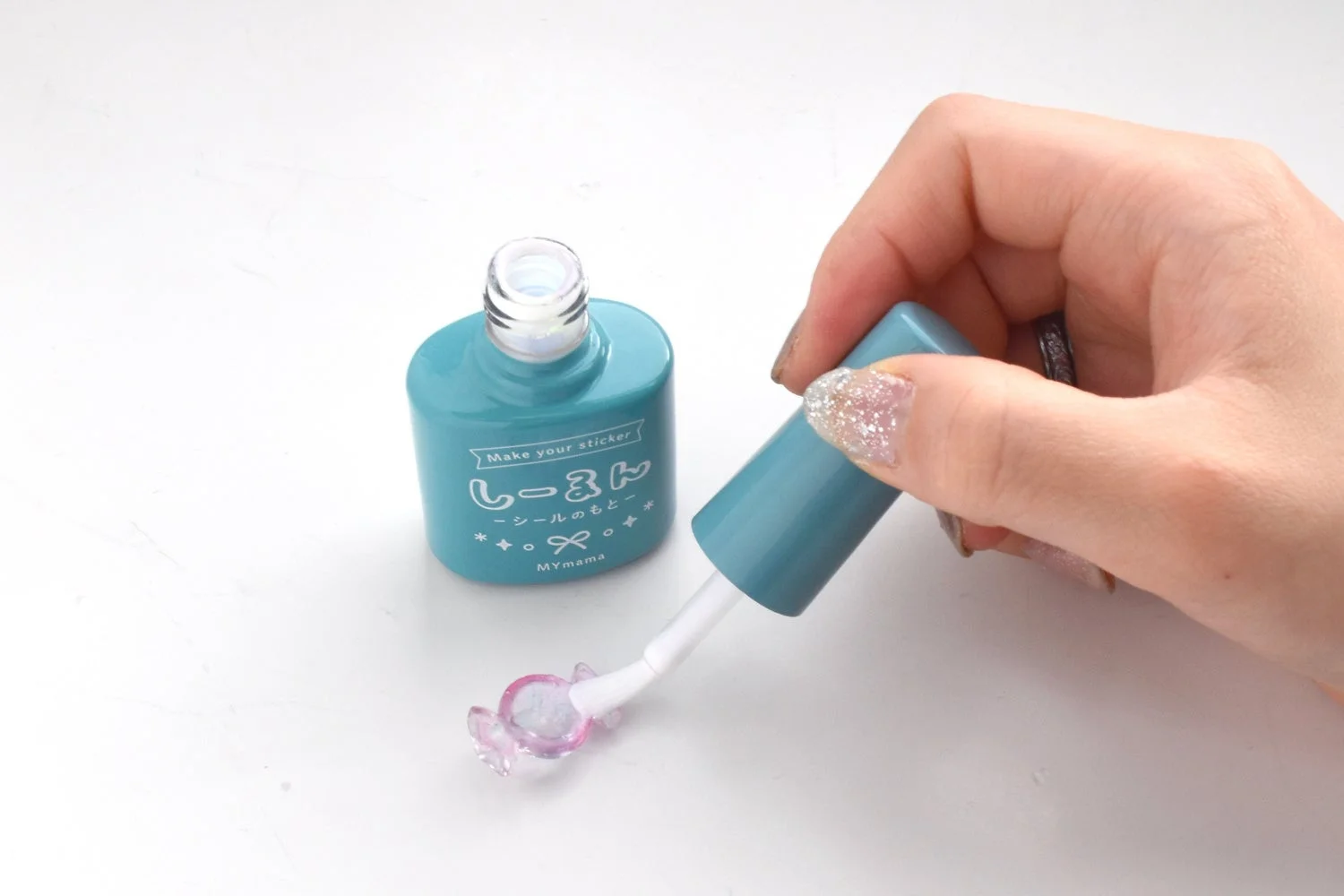 グリッターネイルを施した手が、「しーるのもと」と書かれたターコイズ色のボトルから液体を塗布している様子