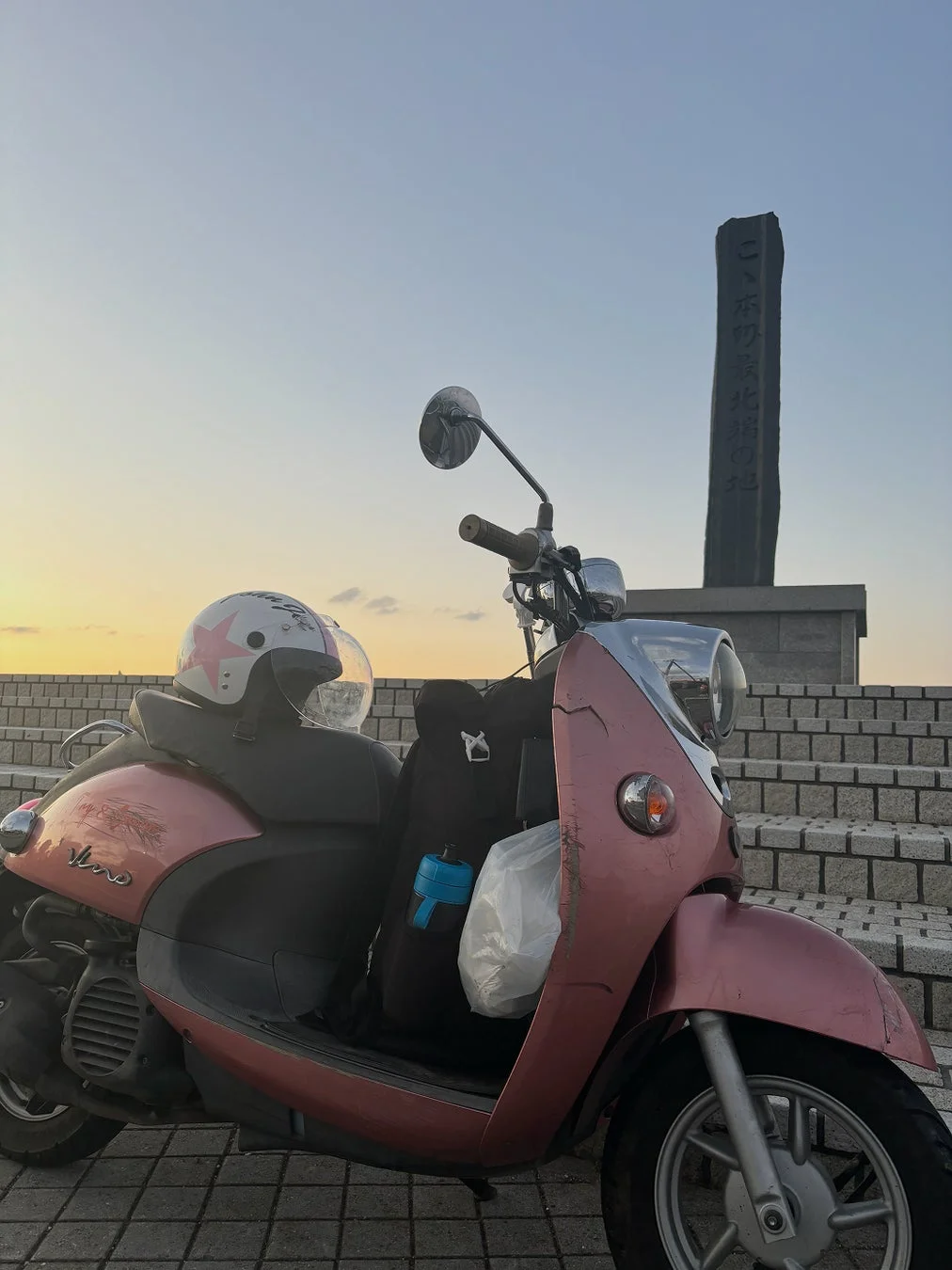夕焼け空の下、日本の最北端の地を示す石碑の前にピンクのスクーターが停まっています。