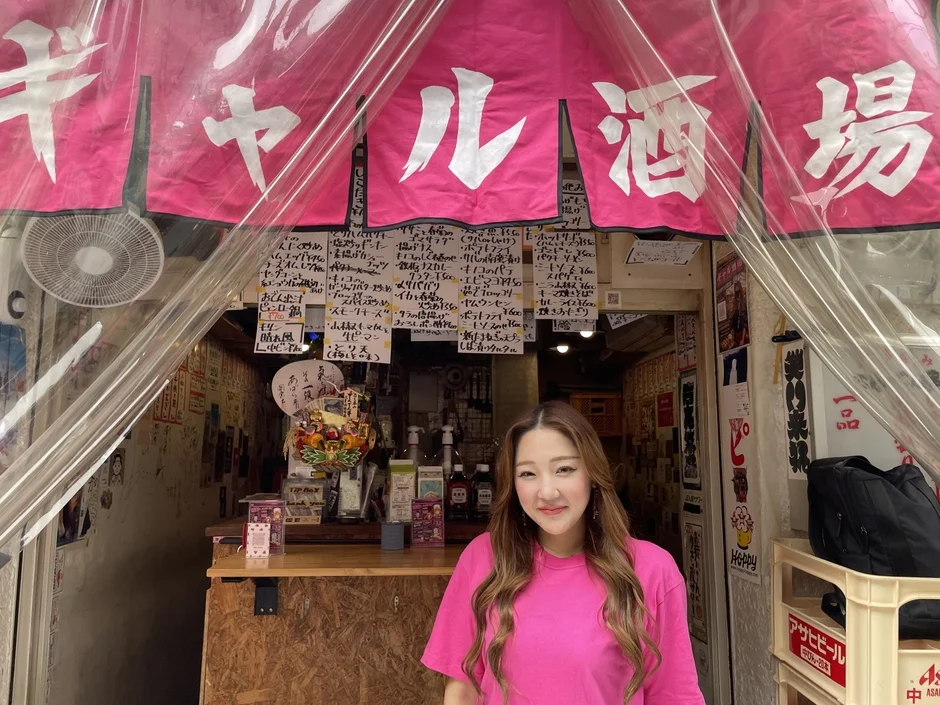 ピンクの暖簾と手書きメニューが特徴的な日本の大衆酒場の前に、笑顔の女性が立っている。