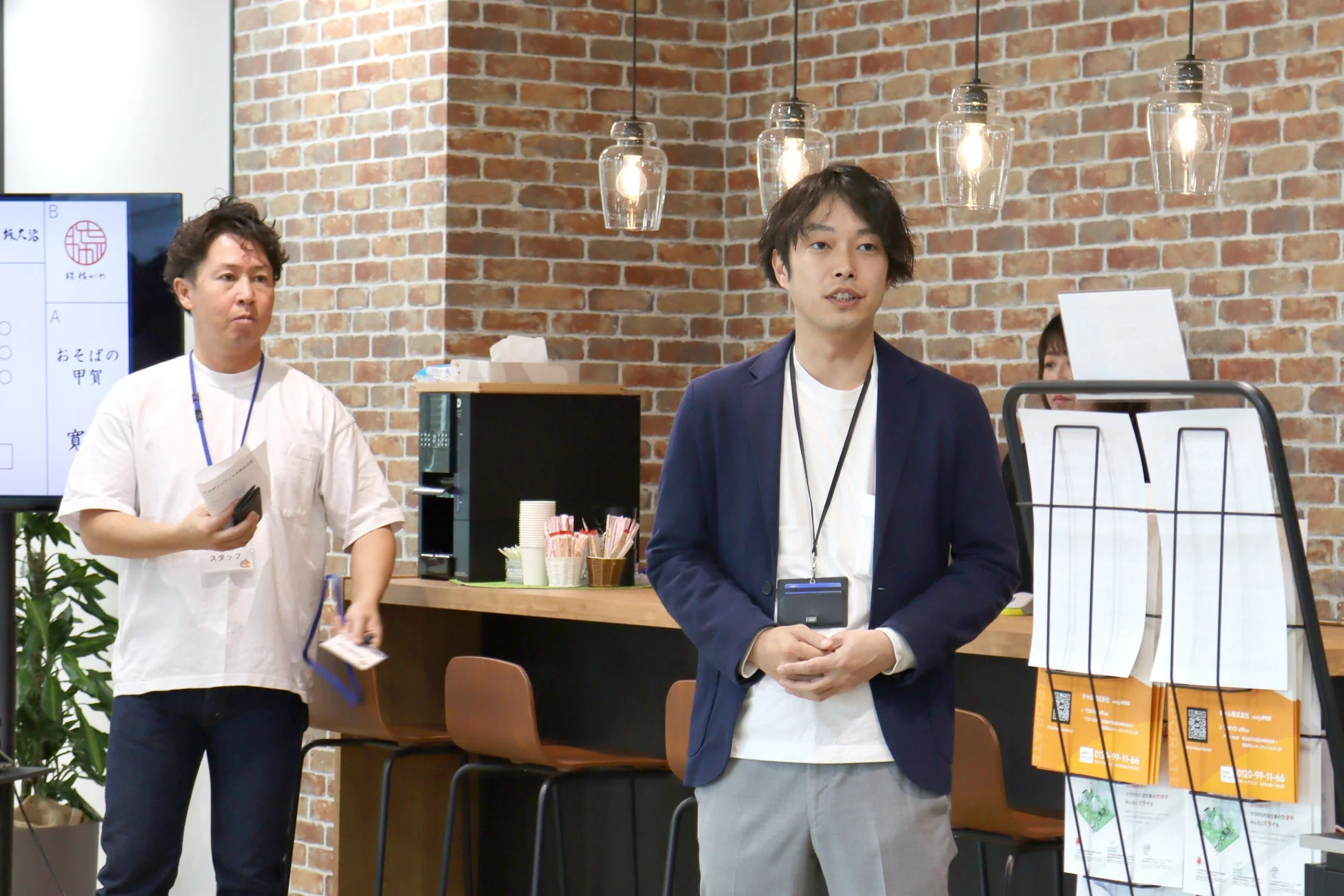 レンガの壁を背景にしたオフィス空間で、2人の男性がプレゼンテーションやイベントに参加している様子です。1人は紺色のジャケットを着て話しており、もう1人は白いTシャツ姿で資料を持っています。