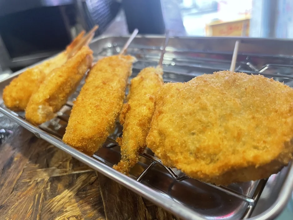 揚げたての串カツやエビフライなど、様々な揚げ物が金属製の網に乗せられて提供されている様子。