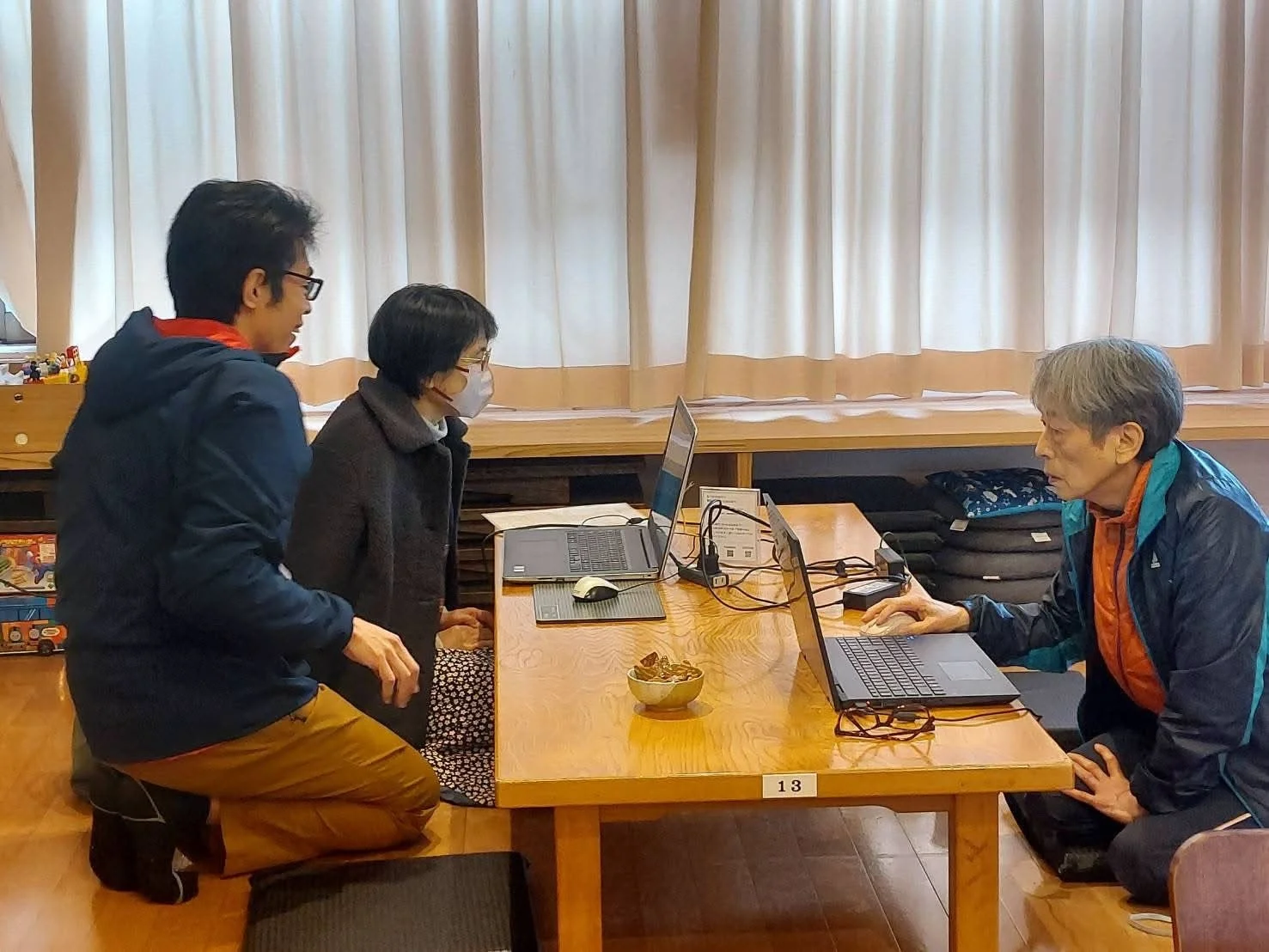 座卓を囲んで3人がノートパソコンを使用している様子。マスクを着用した女性を含め、和やかな雰囲気の中で学習や作業をしているように見えます。デジタル機器を使った勉強会や交流の場面を捉えた一枚です。