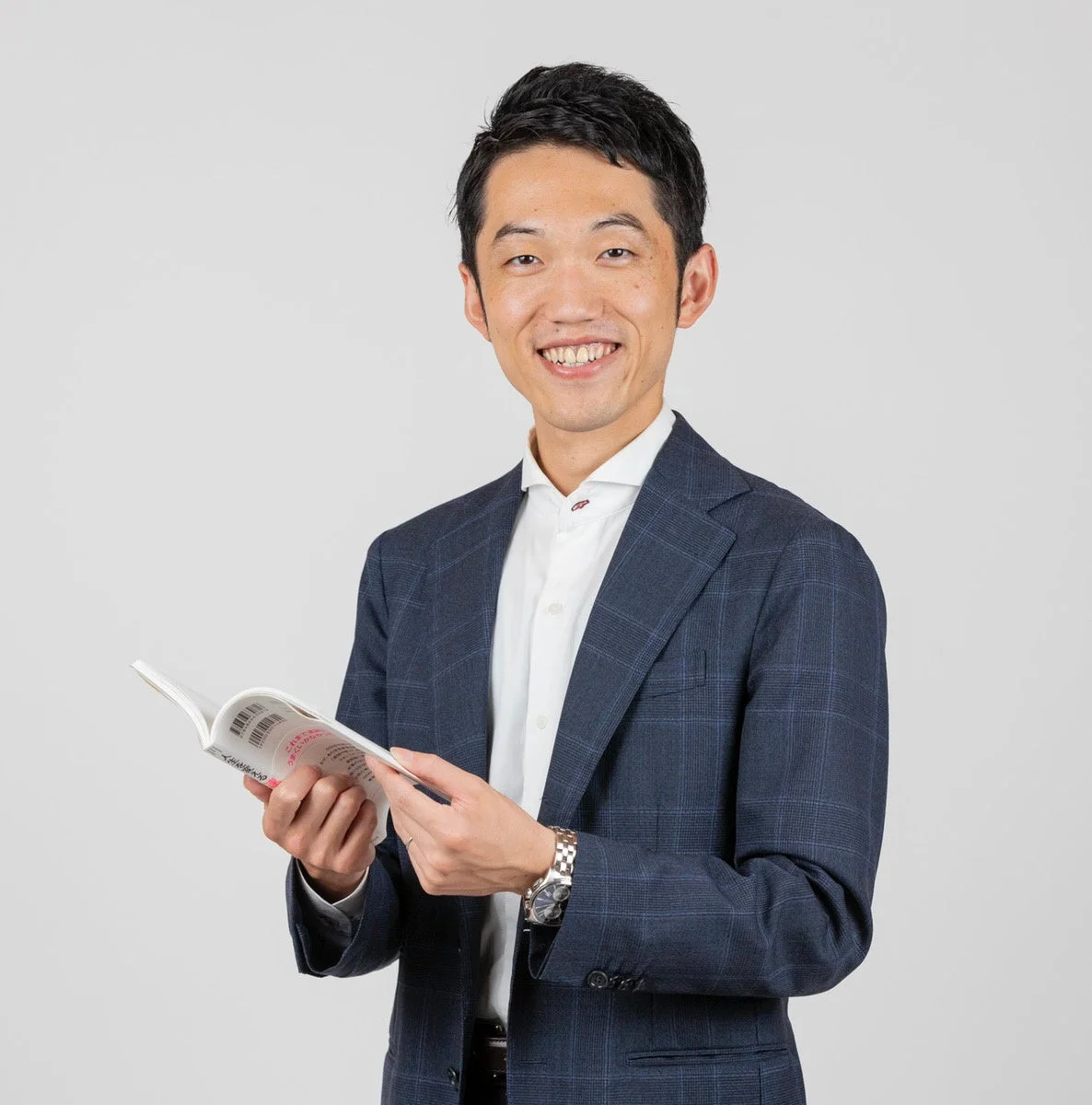 紺色のスーツを着た日本人男性が笑顔で本を持ち、カメラを見ています。清潔感のあるスタジオで撮影されたポートレートです。