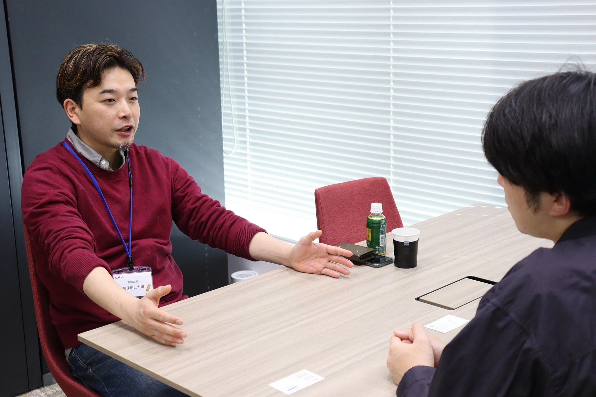 会議室で二人の男性が対話している様子。赤いセーターの男性が身振り手振りを交えて熱心に話しており、ビジネスシーンでの打ち合わせやインタビューの風景を捉えている。