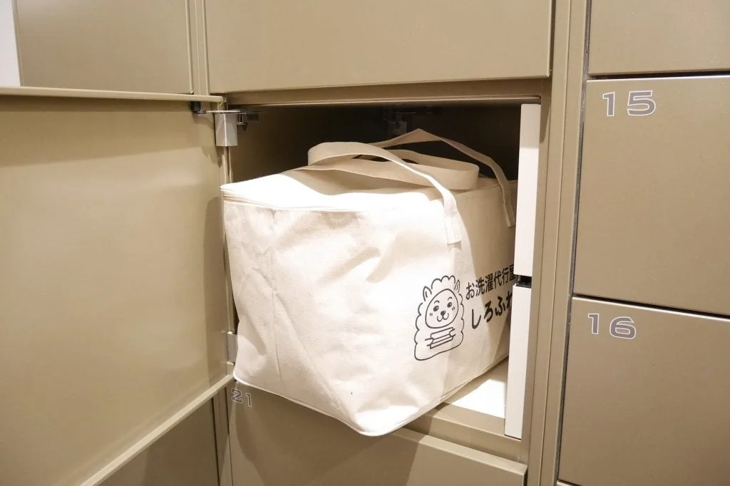 ベージュのロッカーの扉が開き、中に洗濯代行サービス「お洗濯代行屋 しろふわ」のロゴが入った白い布製バッグが収納されている様子