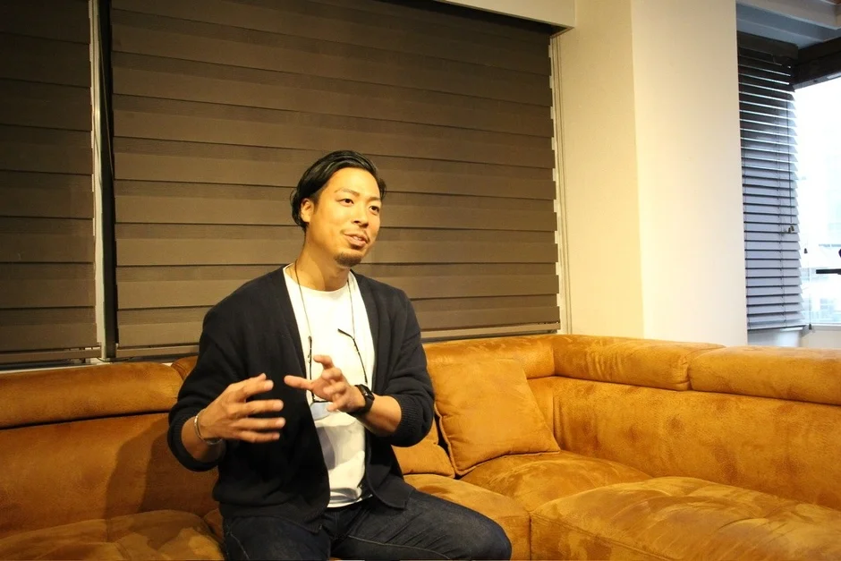 茶色のソファに座った男性が、身振り手振りを交えながら笑顔で話している様子です。