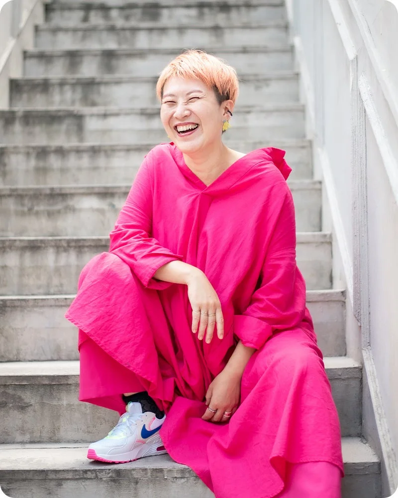 明るいピンクの服を着たショートヘアの女性が、コンクリートの階段に座って楽しそうに笑っています