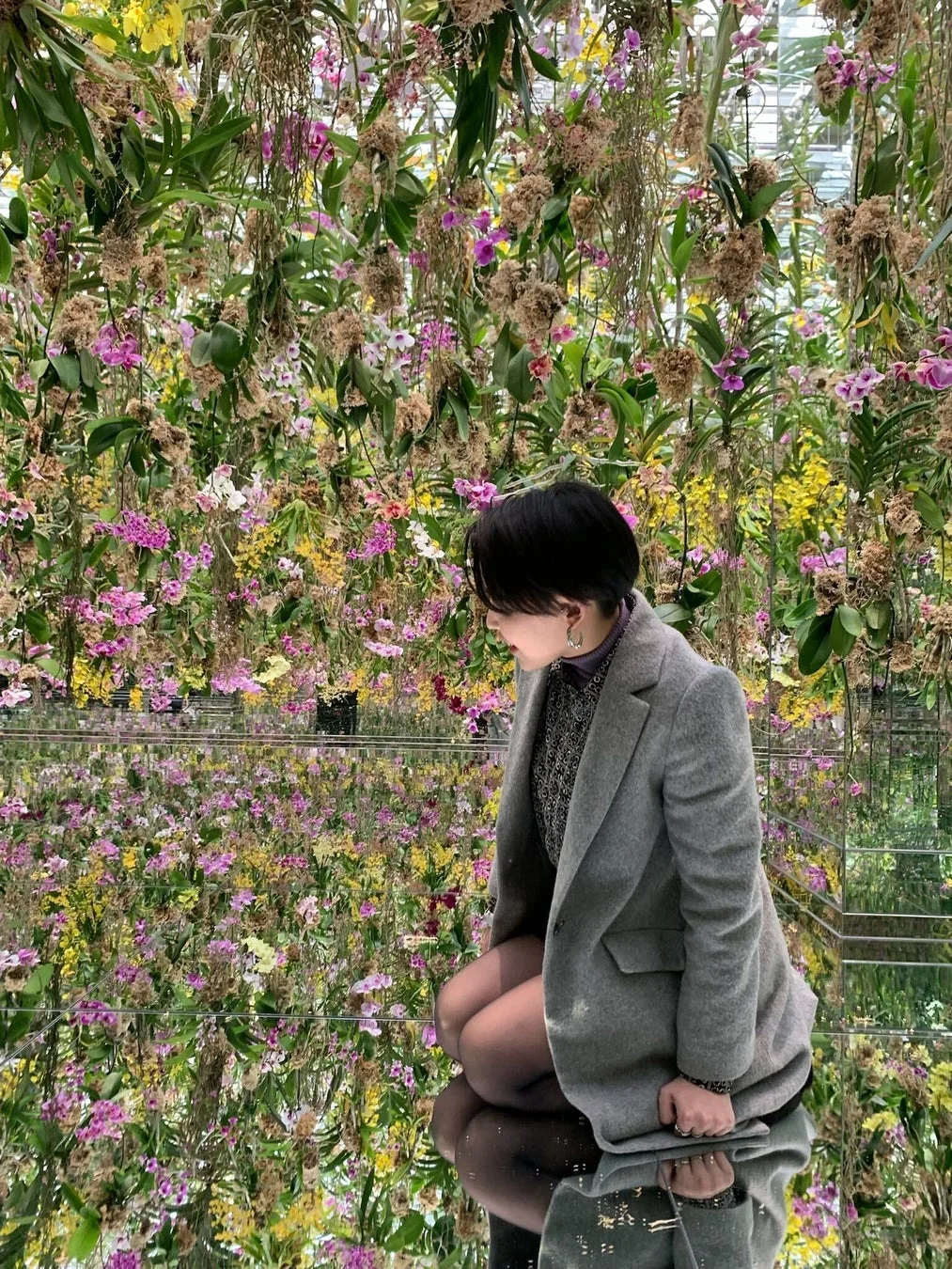 色とりどりの花々が天井から吊り下げられ、床の鏡に映り込む幻想的な空間で、一人の女性がひざまずいています