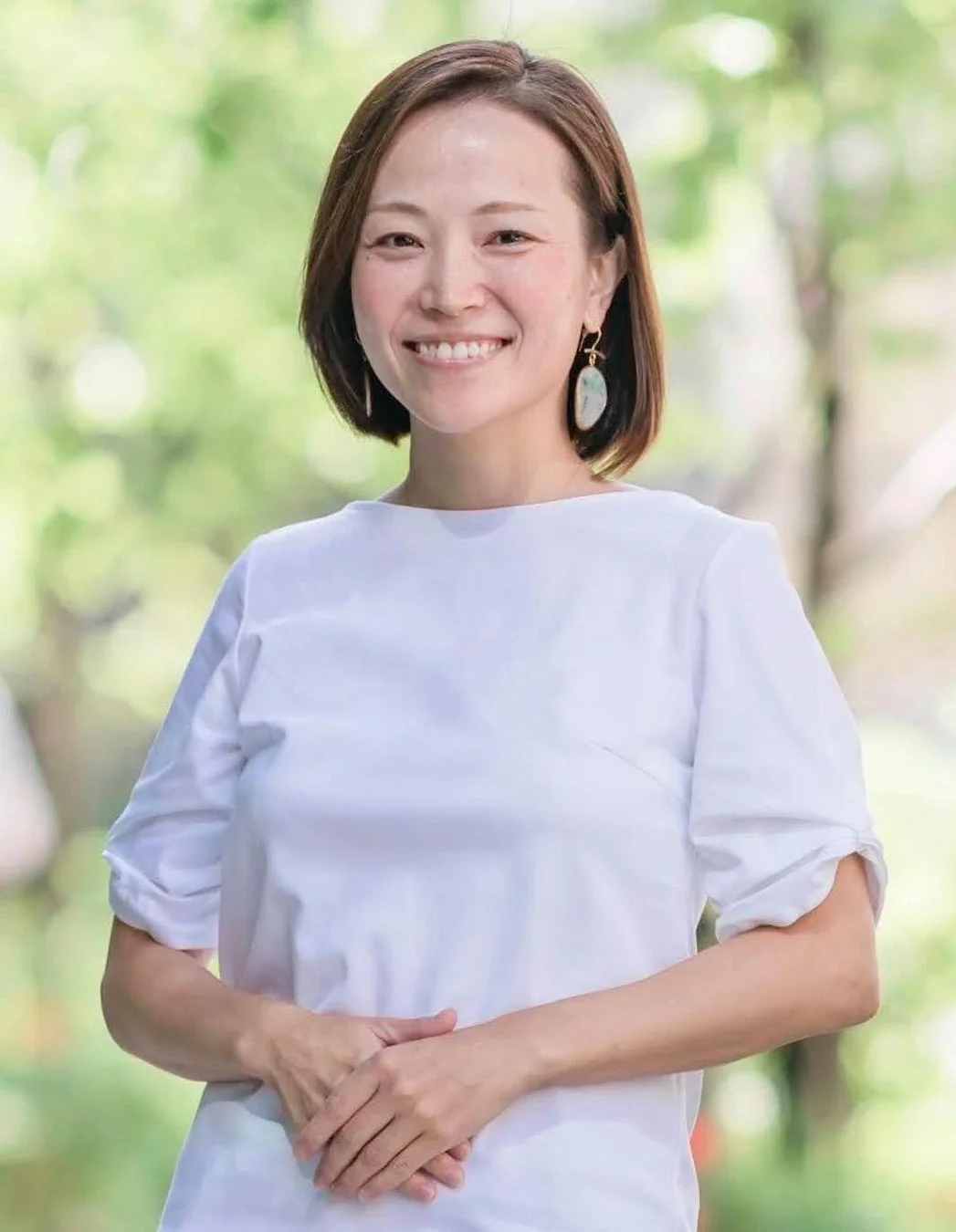 白いトップスを着たショートヘアの女性が、屋外の緑を背景に笑顔でポーズをとっています