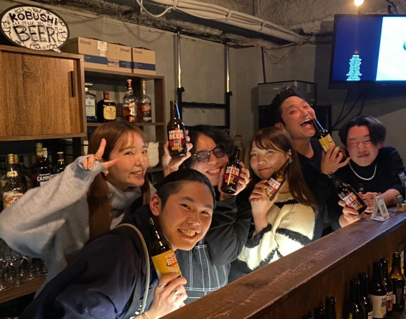 バーカウンターで「KOBUSHI BEER」の瓶ビールを手に笑顔を見せる男女のグループ